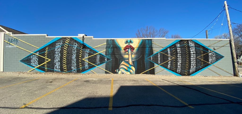Image of mural depicting close up of person's face holding their index finger to their mouth