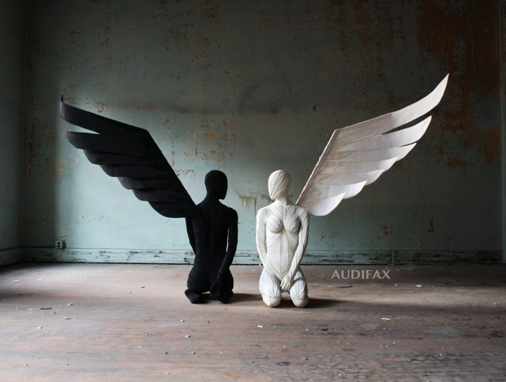 Sculpture of two human-looking figures. One on left is all black. One on right is all white. Both have one wing