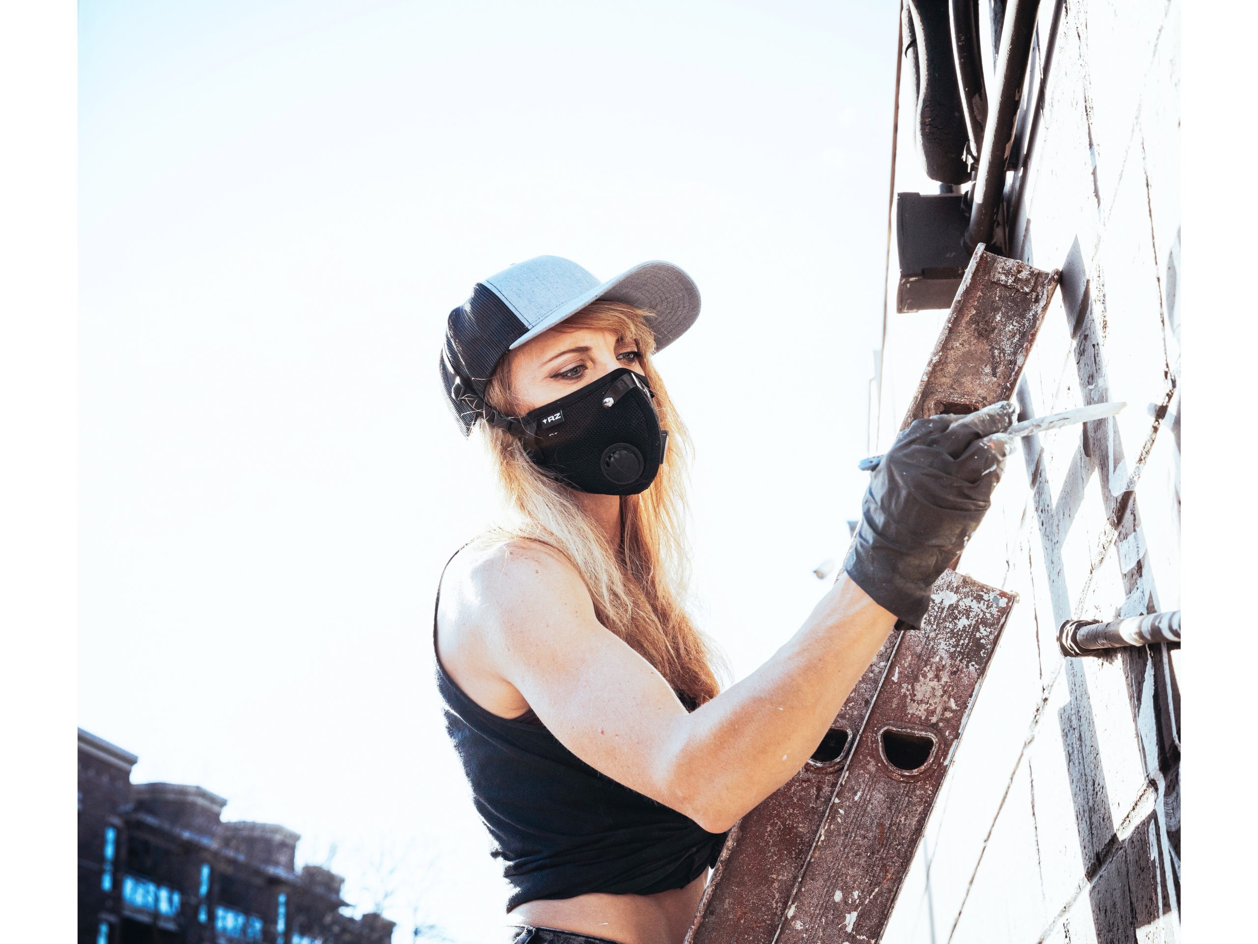 Image of photo of artist wearing a black mask, with long blonde hair and a hat on, working on a mural