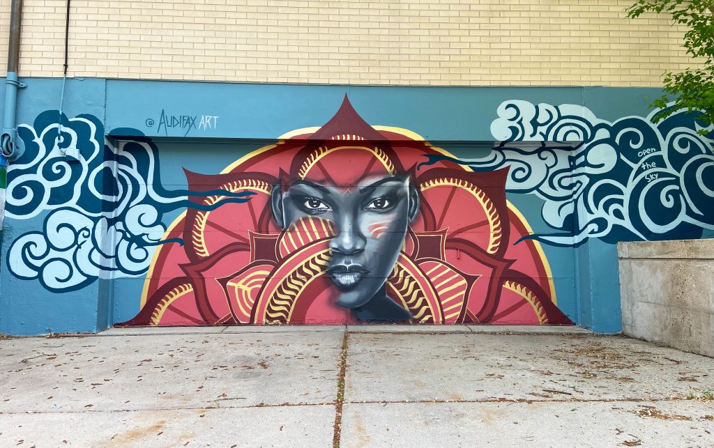 Image of finished mural outside showing a face and red/pink lotus flower behind it; blue swirls on both sides and the text on the right that says 