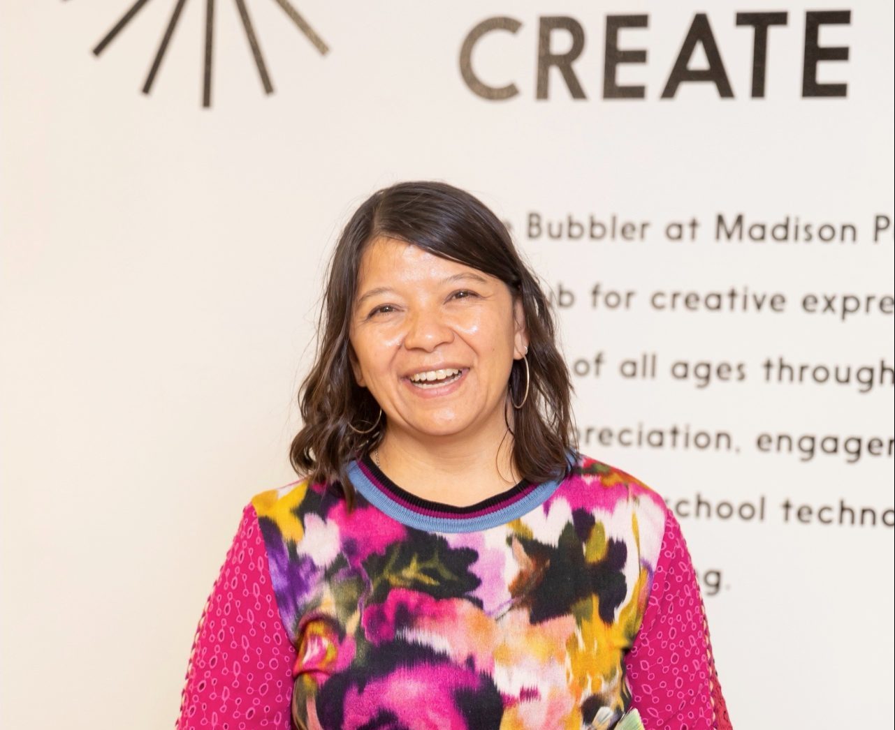 Image of photo of artist standing in front of wall that says "CREATE." Artist is standing and smiling, with long brown hair wearing a colorful sweater and holding paintbrushes