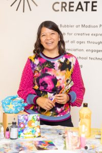 Image of photo of artist standing in front of wall that says "CREATE." Artist is standing and smiling, with long brown hair wearing a colorful sweater and holding paintbrushes