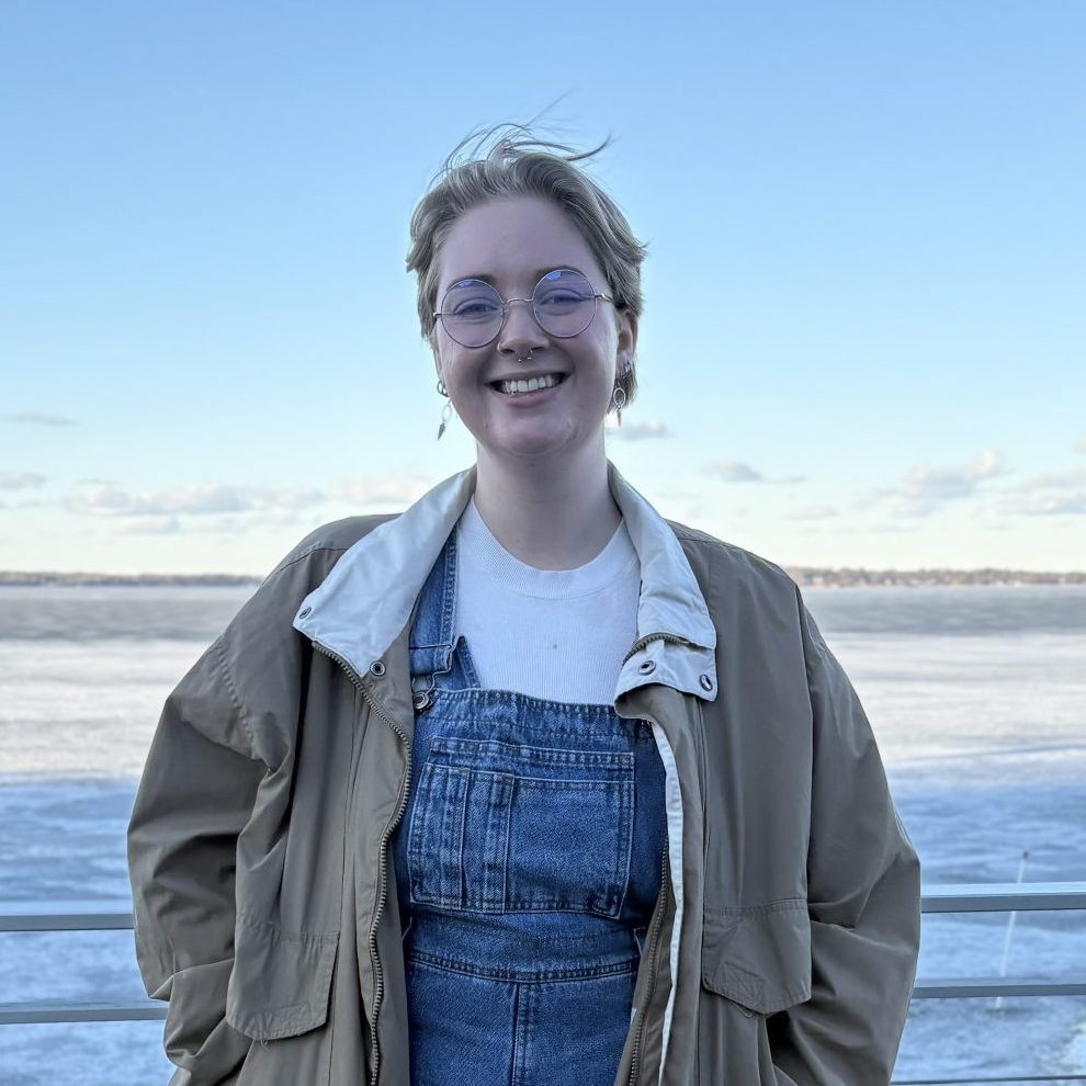 Image of Amelia Stephens. Amelia is wearing denim overalls and a tan jacket and standing in front of a body of water.