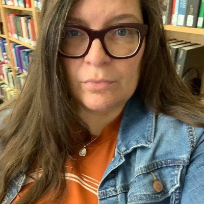 Image of Karla Strand. Karla is wearing glasses, an orange shirt, and a denim jacket. Karla appears in front of bookcases.