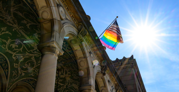 Photograph of American flag and pride flag on top of the Memorial Union at UW-Madison, sunlight burst in sky